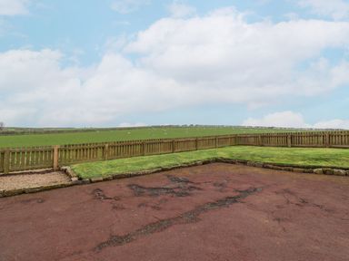 An outdoor area with cracked red pavement grass bordered by stones and a wooden fence with green fields and sheep in the background at Whitestacks in Bamburgh