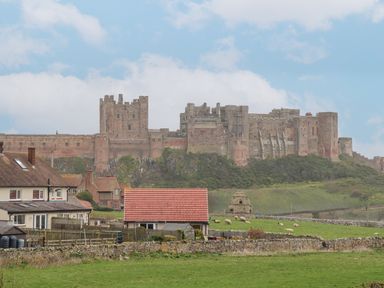 A large stone castle on a hill with houses and green fields with sheep in the foreground at Whitestacks in Bamburgh