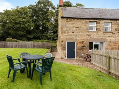 A garden with plastic chairs and a round table beside a stone house with wooden fencing at Bilton Farm Cottage No1 in Alnmouth