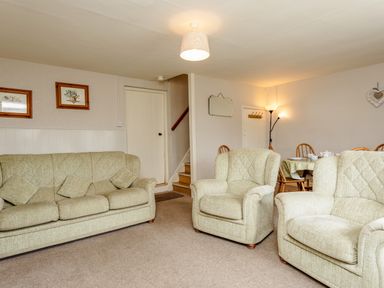 A living room with three upholstered chairs, a dining table with chairs and dishes, two framed pictures on the wall and stairs leading up at Bilton Farm Cottage No1 in Alnmouth