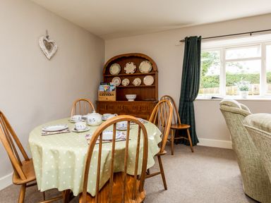 A dining room with a round table covered in a polka dot tablecloth set with tea cups and plates wooden chairs a wooden dresser with plates on display and a window with green curtains at Bilton Farm Cottage No1 Alnmouth