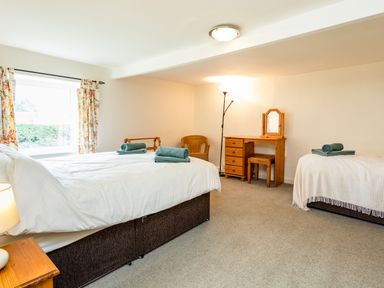 A bedroom with two beds a wooden dressing table a wicker chair and a window with floral curtains at Bilton Farm Cottage No1 in Alnmouth