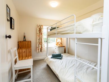 A small bedroom with a white bunk bed a chair wooden steps and a window with floral curtains at Bilton Farm Cottage No1 in Alnmouth