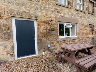 An outdoor area with a wooden picnic table on gravel in front of a stone wall with a blue door and a window with a hanging flower pot at Bilton Farm Cottage No1 in Alnmouth