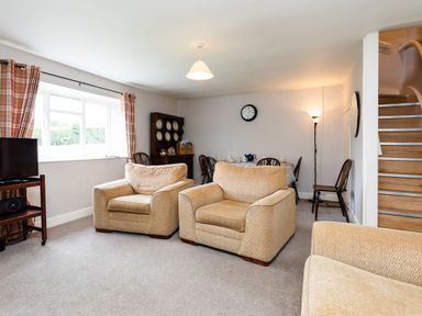 A living room with two beige armchairs a dining table with chairs a wooden cabinet and a staircase at Bilton Farm Cottage No2 in Alnmouth