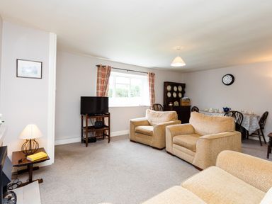 A living room with beige armchairs a television on a stand and a dining table with chairs and plates at Bilton Farm Cottage No2 in Alnmouth