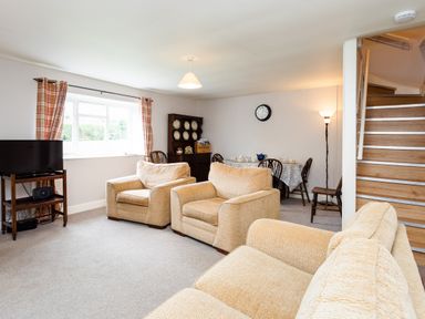 A sitting area with beige armchairs and a television near a window and a dining area with a table and chairs at Bilton Farm Cottage No2 in Alnmouth