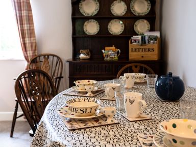 A dining room with a table set with cups bowls and glasses and wooden chairs with a cabinet in the background at Bilton Farm Cottage No2 in Alnmouth