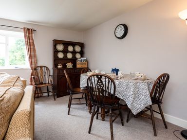 A dining room with a wooden table covered with a cloth and four chairs a wooden cabinet with dishes and a window with plaid curtains at Bilton Farm Cottage No2 in Alnmouth
