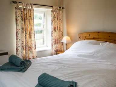 A bedroom with a wooden bedframe white bedding floral curtains a bedside lamp and folded towels on the bed at Bilton Farm Cottage No2 in Alnmouth