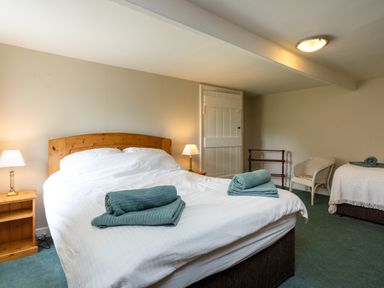 A bedroom with one large bed and one small bed with towels placed on them two bedside tables with lamps a white door and a wicker chair at Bilton Farm Cottage No2 in Alnmouth