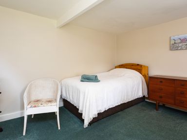 A bedroom with a single bed covered with a white blanket and folded towels a white wicker chair and a wooden dresser with drawers at Bilton Farm Cottage No2 in Alnmouth