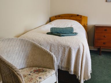 A bedroom with a single bed covered with a white blanket two folded towels on the bed a wicker chair with floral cushion and a wooden chest of drawers at Bilton Farm Cottage No2 in Alnmouth
