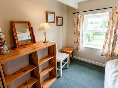 A bedroom corner with a wooden shelving unit with a mirror and lamp a white chair a wooden side table and a window with floral curtains at Bilton Farm Cottage No2 in Alnmouth