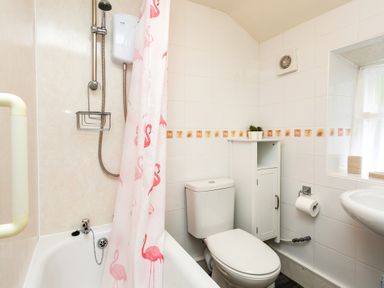 A small bathroom with a bathtub and shower, a toilet, a white cabinet, a window, and a sink at Bilton Farm Cottage No2 in Alnmouth