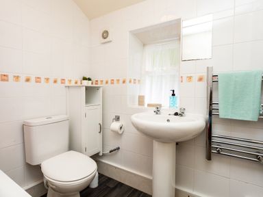 A bathroom with a toilet, pedestal sink, small white cabinet, towel rail with towel, and window at Bilton Farm Cottage No2 in Alnmouth