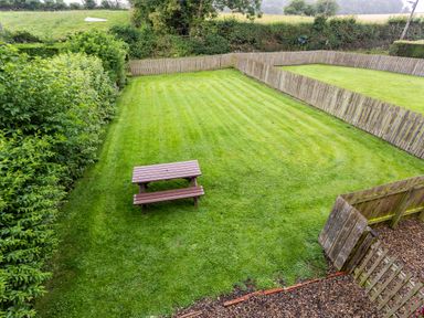 A fenced grass garden with a wooden picnic table and hedge at Bilton Farm Cottage No2 in Alnmouth