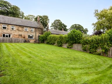 A grassy backyard with a wooden picnic table and a stone house in the background at Bilton Farm Cottage No2 in Alnmouth