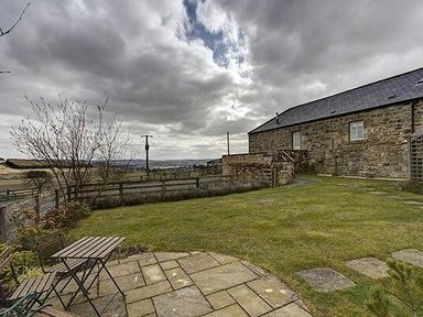 A stone building next to a grassy yard with a small table and chairs on a paved patio at Lantern View in Eglingham