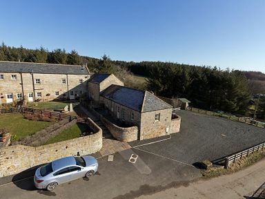 A stone building with a parked silver car and fenced grass area near trees at Lantern View in Eglingham