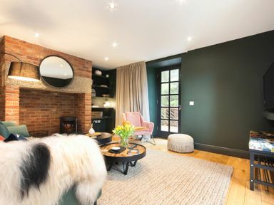 A living room with a brick fireplace a round mirror a pink armchair and a coffee table with flowers at The Bolthole in Eglingham