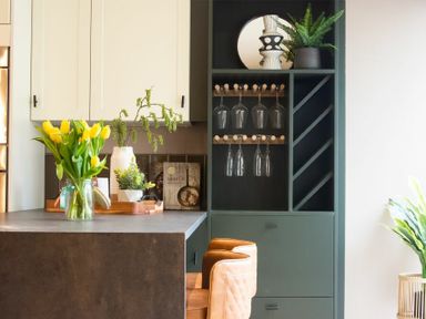 A kitchen corner with a gray countertop a green shelf with wine glasses yellow tulips in a jar and bar stools at The Bolthole in Eglingham