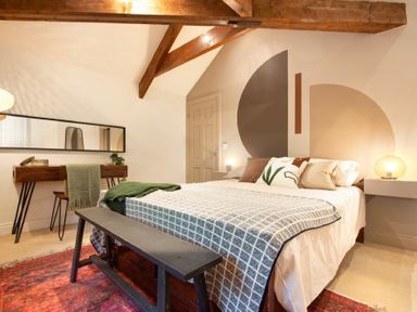 A bedroom with a bed a bench a desk and a mirror under wooden ceiling beams at The Bolthole in Eglingham