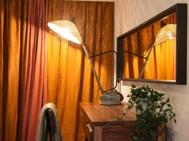 A wooden dresser with a lamp and a plant next to a mirror and orange curtains at The Bolthole in Eglingham