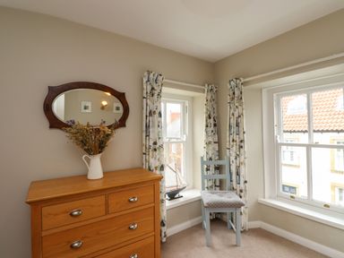 A wooden dresser with a vase of dried flowers and an oval mirror above it next to two windows with patterned curtains and a chair with a cushion in a corner room at Strandline Alnmouth