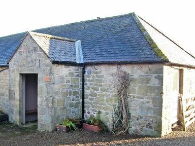 An exterior view of a stone building with a slate roof and an open doorway with plants outside at Swallow Cottage nr Powburn in Powburn