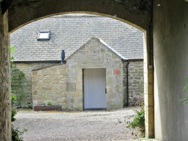 A stone building with a white door seen through a stone archway at Swallow Cottage nr Powburn in Powburn