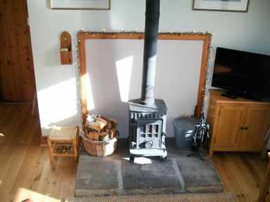 A wood-burning stove with a basket of firewood and a metal ash container next to a wooden cabinet and stool at Swallow Cottage nr Powburn in Powburn