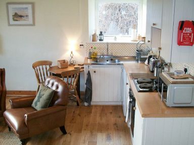 A kitchen with wooden cabinets a sink stove microwave and toaster next to a small dining table with wooden chairs and a brown leather armchair at Swallow Cottage nr Powburn in Powburn