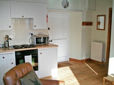 A kitchen area with white cabinets a microwave a stove and oven a brown leather chair and a wall clock at Swallow Cottage nr Powburn in Powburn