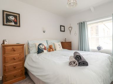 A bedroom with a bed pillows a teddy bear and rolled towels two wooden bedside cabinets and a window with curtains at Aln Cottage Alnwick