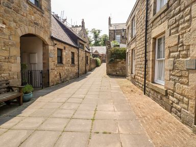 A paved narrow street with stone buildings on both sides and a wooden bench on the left at Aln Cottage Alnwick