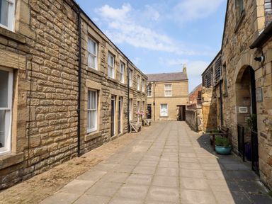A paved alleyway with stone buildings on both sides and benches with potted plants at Aln Cottage Alnwick