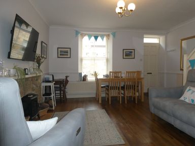 A living room with sofas a dining table wooden chairs a wall-mounted tv a fireplace and a window with bunting at Aln Cottage Alnwick