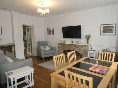 A living room with wooden dining table and chairs a stone fireplace with a mounted TV and gray armchairs at Aln Cottage in Alnwick