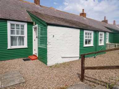 A green and white cottage with multiple windows and a white door on a gravel yard at Armstrong Cottages No2 in Bamburgh