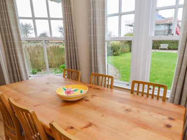 A dining area with a wooden table and chairs by large windows overlooking a garden at Armstrong Cottages No2 Bamburgh