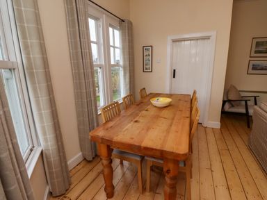 A dining area with a wooden table and six chairs near windows with beige curtains at Armstrong Cottages No2 in Bamburgh