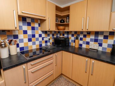 A kitchen corner with wooden cabinets a black countertop a stovetop a microwave and cupboards at Armstrong Cottages No2 Bamburgh