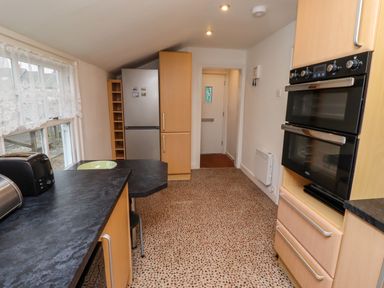 A kitchen with black countertops wooden cabinets a double oven a refrigerator and a window with lace curtains at Armstrong Cottages No2 Bamburgh