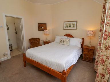 A bedroom with a wooden bed and white bedding two bedside tables with lamps a brown armchair and floral curtains at Armstrong Cottages No2 in Bamburgh