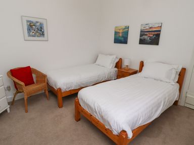 A bedroom with two single beds a wicker chair and paintings on the walls at Armstrong Cottages No2 in Bamburgh
