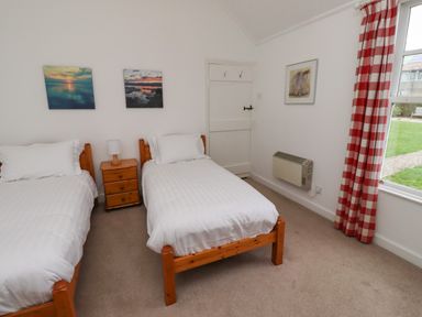 A bedroom with two single beds a wooden nightstand between them two paintings on the wall a window with checkered red and white curtains and a heating unit on the wall at Armstrong Cottages No2 in Bamburgh