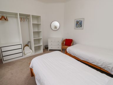 A bedroom with two single beds white shelving unit with hangers white chest of drawers wicker chair with red cushion at Armstrong Cottages No2 in Bamburgh