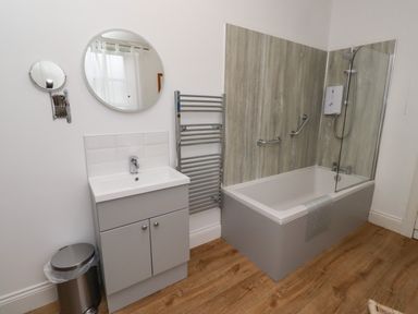 A bathroom with a bathtub with shower and glass screen a sink with cabinet two mirrors a towel radiator and a trash bin at Armstrong Cottages No2 Bamburgh