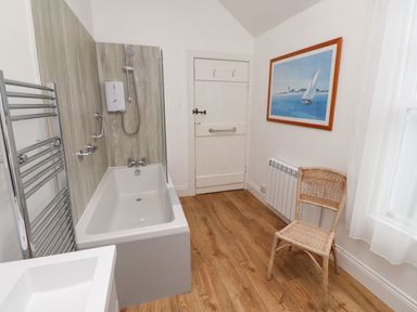 A bathroom with a bathtub shower a wicker chair a radiator a painting on the wall and wooden flooring at Armstrong Cottages No2 in Bamburgh